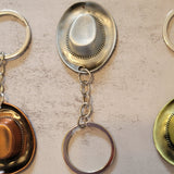 Three cowboy hat keychains arranged side by side on a light beige textured surface. The keychains feature vintage-style metal hat emblems with attached key rings: one is gold-toned, one is silver-toned, and one is green-toned.