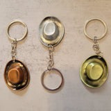Three cowboy hat keychains arranged side by side on a light beige textured surface. The keychains feature vintage-style metal hat emblems with attached key rings: one is gold-toned, one is silver-toned, and one is green-toned.