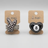 Two small jewelry pieces on display cards against a light textured background. The left item is a silver-toned ring with a black-and-white checkered bunny head and a wrapped band.