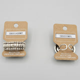 Two small metal keyring accessories on beige display cards, photographed on a textured beige surface. One item has a silver key ring and the other has a coiled silver metal component.