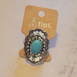 A silver-toned filigree ring with a turquoise oval center stone, displayed on a tan cardboard hanger labeled 'tipi.' against a light textured surface.